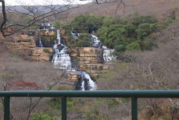 Una impresionante cascada de varios niveles fluye sobre acantilados rocosos, rodeada de árboles secos y follaje verde, vista desde detrás de una barandilla verde en un paisaje sereno.