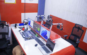 Se trata de un moderno estudio de radio con micrófonos, auriculares, mezclador de audio, pantallas de ordenador y sillas ergonómicas. Los colores son rojo, blanco y azul.