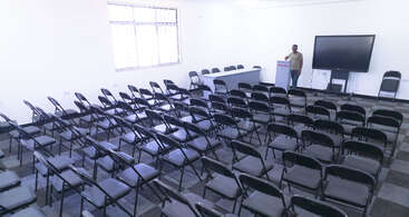 The image shows an empty conference room with many neatly arranged black chairs. One person stands at a podium near a large screen and a long table.
