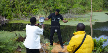 Trois hommes sont en plein air au bord d'un étang, entouré de verdure. L'un prend la pose, l'autre filme avec un appareil photo, tandis que le troisième observe en veste jaune.