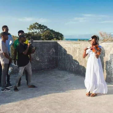 Una mujer vestida de blanco toca el violín en una azotea de hormigón, con un grupo de personas observándola y un fotógrafo tomando imágenes.