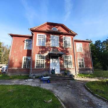 Une grande maison ancienne en bois rouge se dresse sous un ciel bleu éclatant, avec sa peinture écaillée, ses nombreuses fenêtres, son porche à l'avant et sa brouette bleue à l'extérieur.