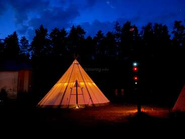 Une tente tipi lumineuse se détache dans la nuit, éclairée dans la forêt sombre. Une lumière chaude s'en échappe, tandis que des lumières colorées en arrière-plan rehaussent la tranquillité de la scène.