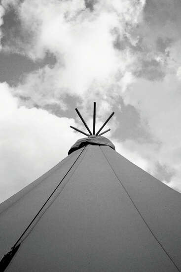 This black and white photo shows the top of a tipi against a cloudy sky, focusing upward to highlight its poles and the fabric structure.