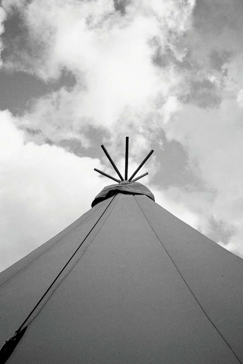 Cette photo en noir et blanc montre le sommet d'un tipi contre un ciel nuageux, avec un cadrage vers le haut pour mettre en valeur ses poteaux et la structure en tissu.