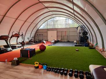 Dieses Bild zeigt ein geräumiges Fitnessstudio mit Laufbändern, Ellipsentrainern, Gewichten, Kettlebells, einem großen Gymnastikball, Kunstrasen, Matten und verschiedenen Trainingsgeräten in einem Kuppelbau.