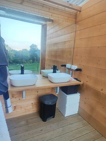 Das ist ein modernes, rustikales Badezimmer mit Holzwänden, Doppelwaschbecken, schwarzen Wasserhähnen, einem schwarzen Mülleimer, einem offenen Fenster, weißen Schubladen und einem schönen Blick nach draußen.