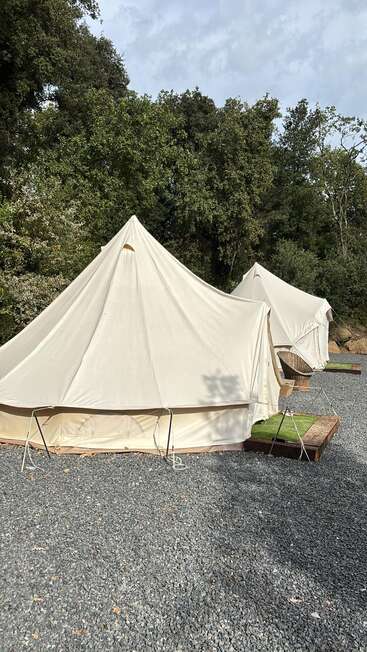 Zwei große, cremefarbene Zelte stehen auf Kiesflächen, umgeben von üppig grünen Bäumen und einem teilweise bewölkten Himmel, der eine friedliche, rustikale Campingszene schafft.