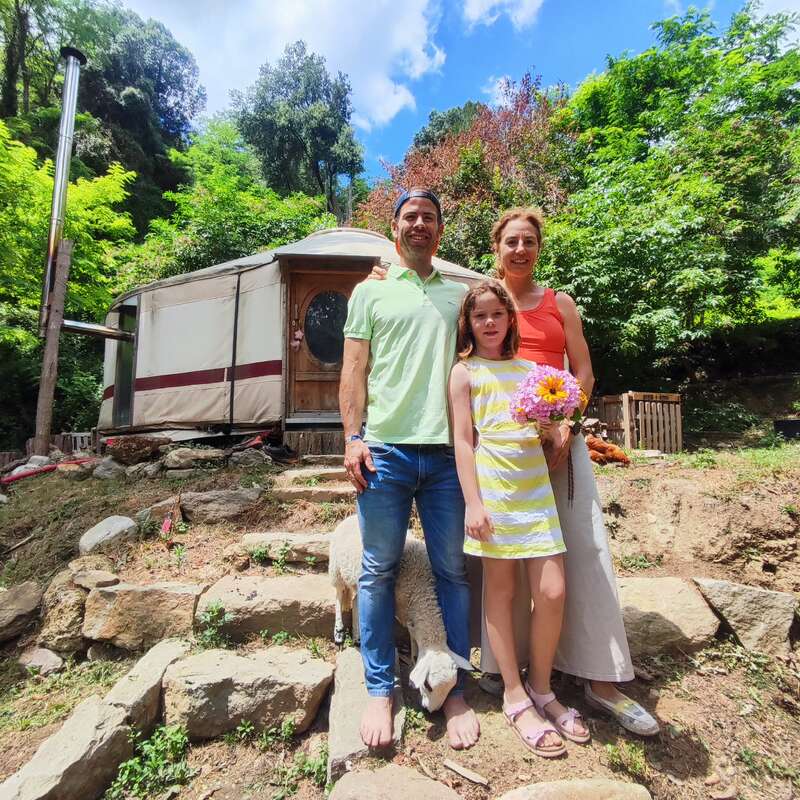 Eine lächelnde dreiköpfige Familie steht barfuß auf Steinstufen vor einer Jurte, umgeben von üppigem Grün, mit einem Schaf und einem Blumenstrauß.