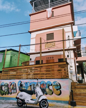 Hay una moto blanca aparcada delante de un colorido mural en el que se lee «CASA SANTURCE», con un edificio rosa y un cielo azul al fondo.