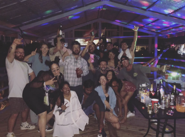 Un animado grupo de amigos en una colorida fiesta en una azotea, posando y sonriendo, rodeados de bebidas, luces y adornos. Parece que todos se lo están pasando en grande.