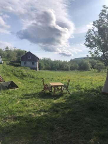 Uma cena campestre tranquila com uma pequena mesa e duas cadeiras sobre a grama verde, uma casa rústica ao fundo e nuvens fofas e dramáticas no alto.