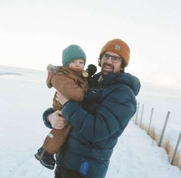 Um homem segurando uma criança pequena está do lado de fora em uma paisagem com neve. Ambos estão vestidos com roupas de inverno, sorrindo e aproveitando o dia frio e claro.