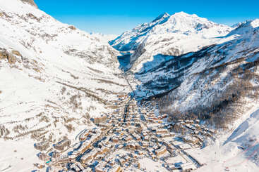 La imagen representa una serena ciudad de montaña enclavada en un valle, con edificios y árboles cubiertos de nieve, sobre un fondo de majestuosas montañas nevadas bajo un cielo azul despejado.