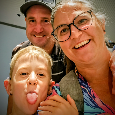 A joyful family selfie features a smiling woman with glasses, a cheerful man in a cap, and a playful child sticking out their tongue.