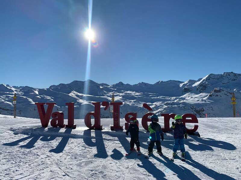 La imagen muestra a tres niños delante de un gran cartel "Val d'Isère" en la ladera de una montaña nevada, con el sol brillando en lo alto.