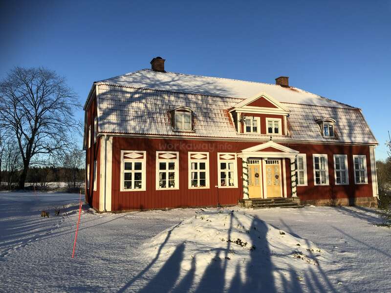 The image depicts a two-story red house with white trim and a snow-covered roof, featuring multiple windows and a yellow door, set against a clear blue sky.