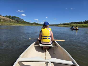Une personne est assise dans un canoë, vêtue d'un gilet de sauvetage jaune et d'une casquette bleue, pagayant sur une rivière calme sous un ciel bleu clair, avec d'autres personnes à proximité.
