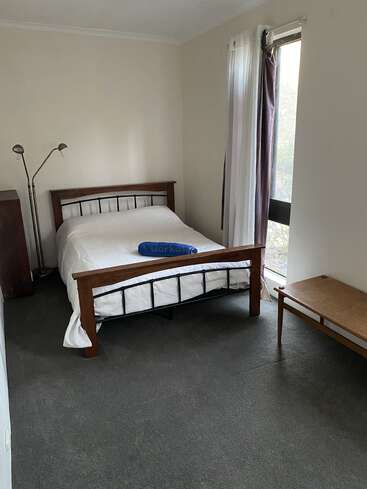 Ein einfaches Schlafzimmer mit einem Holzbett, weißem Bettzeug, einem blauen Kissen, einer Stehlampe, einer Holzbank, einem großen Fenster, weißen Vorhängen und hellen Wänden. Minimalistische Einrichtung.