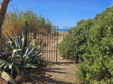 Un portail rouillé ouvert mène à un chemin de sable, entouré de buissons verts et d'agaves, qui laisse entrevoir le bleu de la mer et du ciel.