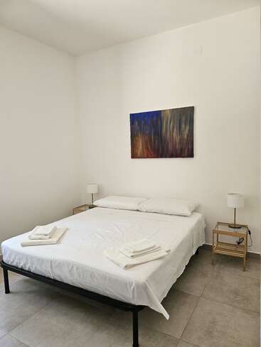 Esta imagen muestra un dormitorio minimalista con una cama bien hecha, sábanas blancas, dos mesillas de noche, lámparas, toallas apiladas y obras de arte abstracto en la pared.