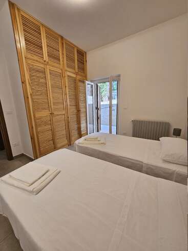 Esta imagen muestra un dormitorio ordenado con dos camas individuales, ropa de cama blanca, toallas dobladas, un gran armario de madera, una puerta abierta a un patio y una decoración neutra.