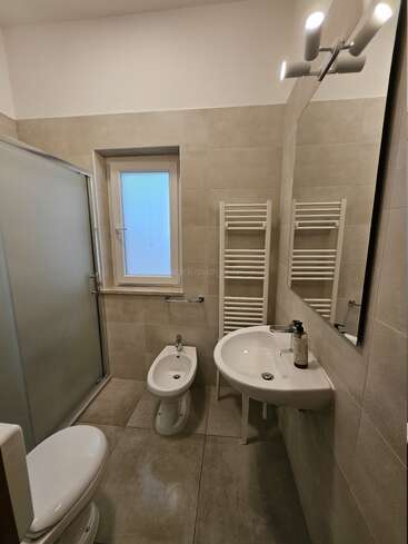 Se trata de un moderno cuarto de baño con azulejos de color beige, que contiene un inodoro, un bidé, un lavabo con espejo, una cabina de ducha, una ventana, un radiador toallero y una elegante iluminación de pared.