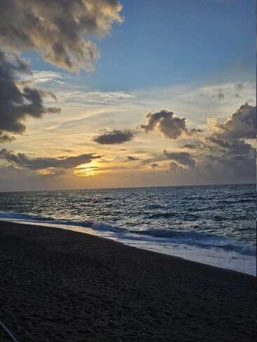 La luz dorada del sol se asoma a través de las espectaculares nubes sobre el océano. Las suaves olas se encuentran con la playa de arena oscura, creando un ambiente tranquilo y apacible bajo un cielo azul que se desvanece.