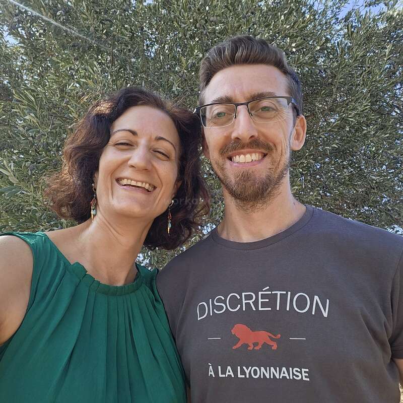 Une femme souriante vêtue d'une robe verte et un homme portant des lunettes et un T-shirt imprimé prennent un selfie joyeux en plein air, avec des arbres feuillus en arrière-plan.