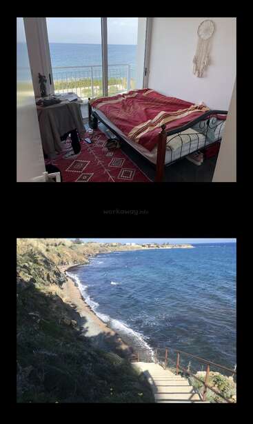 A cozy bedroom with a sea view, red bedding, and a dreamcatcher. Below, stairs lead down to a serene, rocky coastline and clear blue ocean.