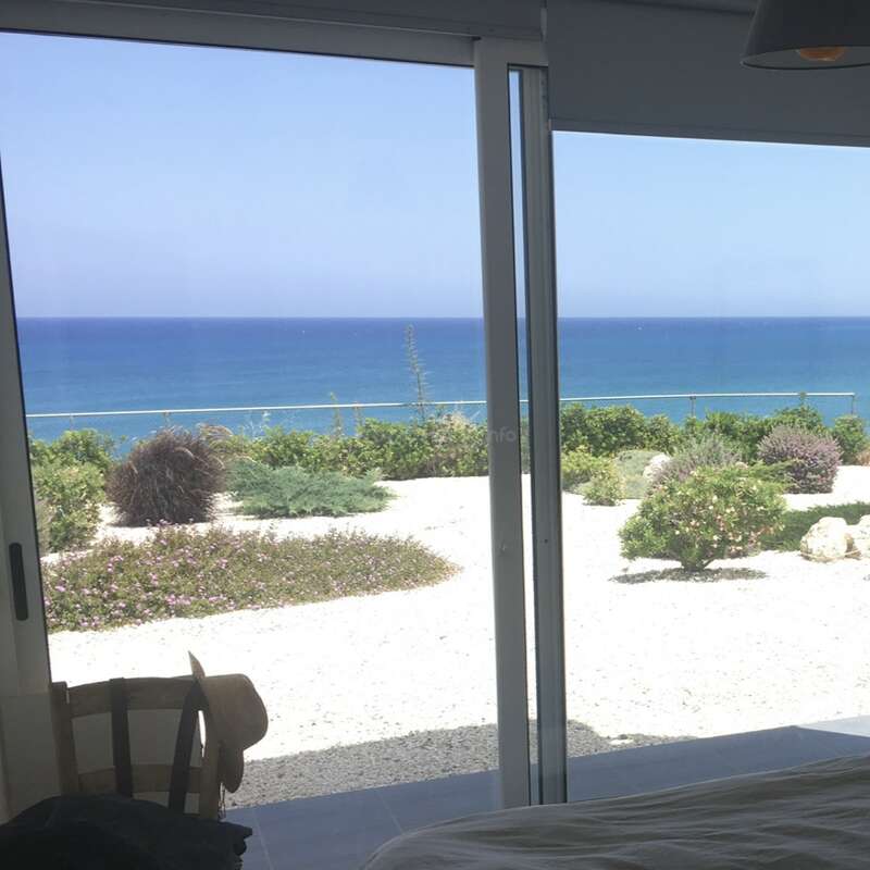 A serene view through glass doors reveals a beautiful garden with green bushes, sandy ground, and a vast blue ocean under a clear sky. Peaceful setting.