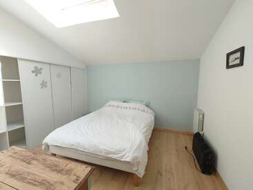 This image shows a simple, bright bedroom with a skylight, light blue accent wall, a double bed, white bedding, wardrobe, heater, and a wooden table.