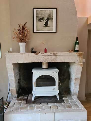 Une cheminée rustique et confortable avec un poêle blanc vintage, un foyer en pierre et un manteau décoré d'un vase de fleurs séchées, d'une bouteille et d'une photographie encadrée en noir et blanc.