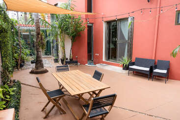 This image shows a cozy outdoor patio with a wooden table, black chairs, greenery, string lights, tropical plants, a red wall, and lounge seating.