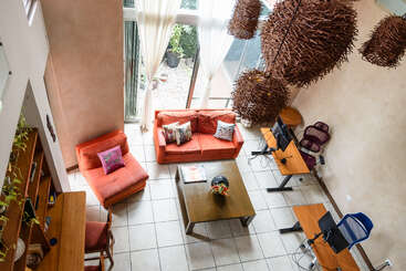 This modern living room features bright orange furniture, a wooden coffee table, workstations with computers, large windows with sheer curtains, hanging twig lamps, and tiled flooring.