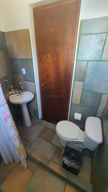 This image shows a small bathroom with tiled walls and floor, a white toilet, pedestal sink, a wooden door, and a shower curtain. A trash bin is nearby.