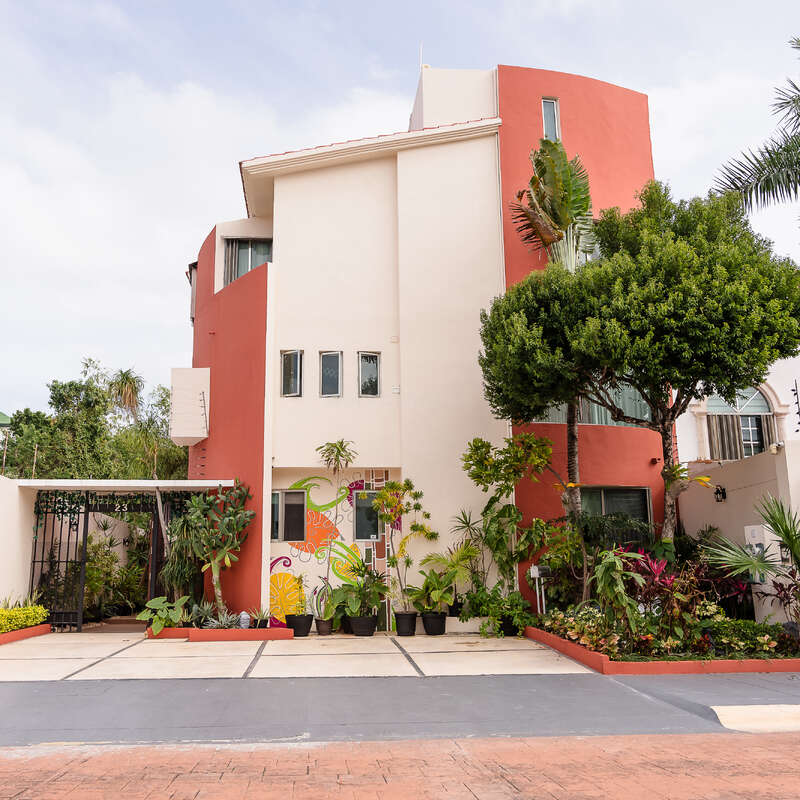 A modern, three-story house with white and terracotta walls, numerous windows, vibrant mural, lush green plants, and well-maintained landscaping, set under a bright sky.