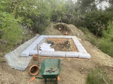 This image shows a construction site in a forested area, with sandbags forming a square foundation, a wheelbarrow, tools, drainage pipes, and scattered gardening supplies.