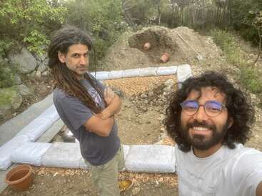 Two men stand outdoors at a construction site, surrounded by trees. They are smiling, with bags and gravel forming the foundation of an earthbag structure behind them.