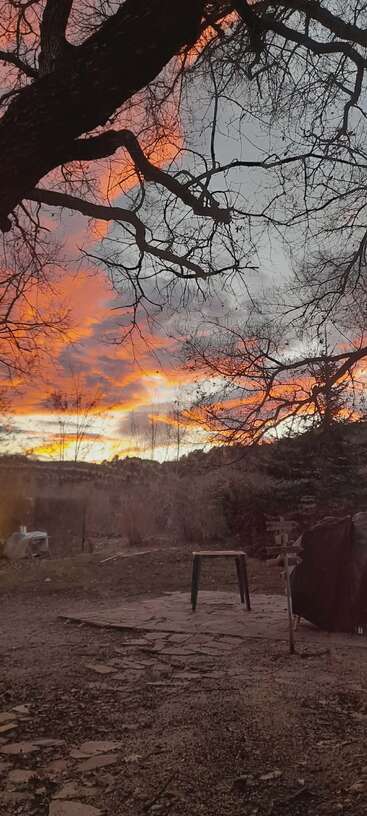 Un coucher de soleil éclatant peint le ciel de teintes orange et rose. Les branches dénudées des arbres encadrent la cour tranquille, parsemée de meubles et d'allées en pierre naturelle.