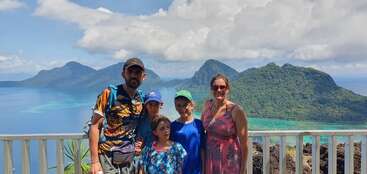 Une famille de cinq personnes pose sur un point de vue panoramique. Derrière eux, des îles verdoyantes, des eaux turquoise et un ciel partiellement nuageux créent une atmosphère de paradis tropical.