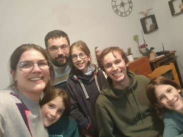 Une famille heureuse de six personnes pose ensemble à l'intérieur, souriant à l'appareil photo. L'arrière-plan comprend un décor de maison, une horloge murale, des plantes et une armoire en bois.