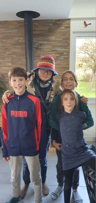 Quatro crianças, sorrindo e posando em um ambiente fechado perto de uma janela. Uma delas usa um chapéu colorido e óculos. Suas roupas são casuais. A luz natural ilumina o cômodo aconchegante.