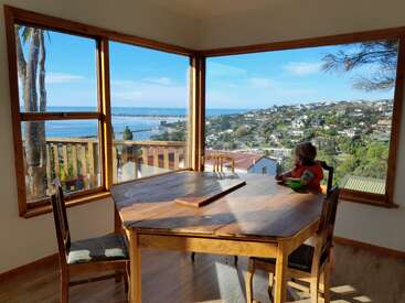 A child sits at a wooden dining table by large windows, enjoying a bowl, with stunning ocean and hillside views under a bright blue sky.