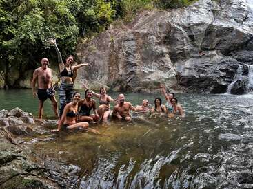 A group of people pose for a photo in a river, with rocks and trees in the background, surrounded by lush greenery.
