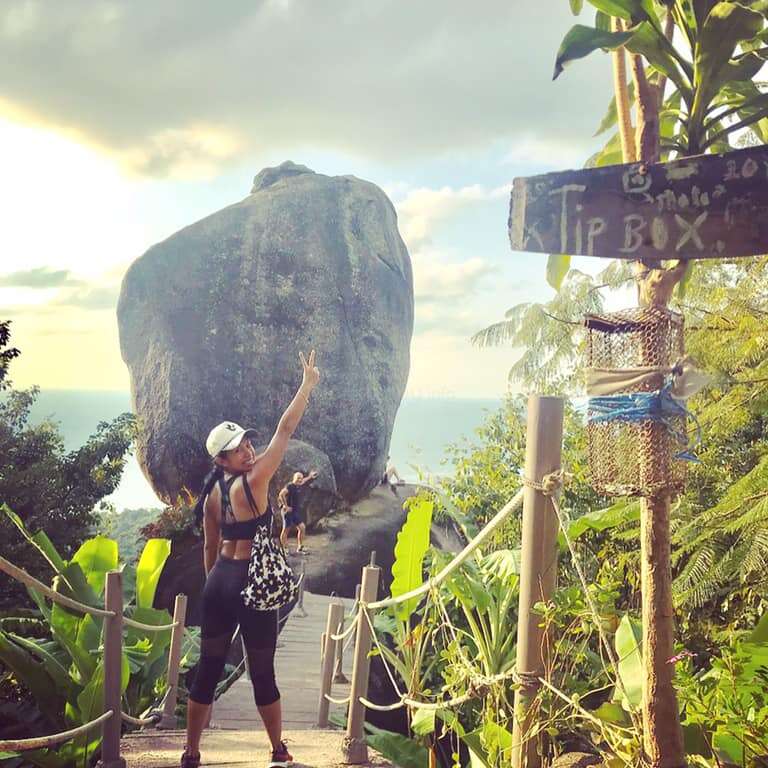 A woman stands on a wooden walkway, posing with her left arm raised, in front of a large rock formation, with lush greenery and a sign that reads \"Tip Box\" in the background.