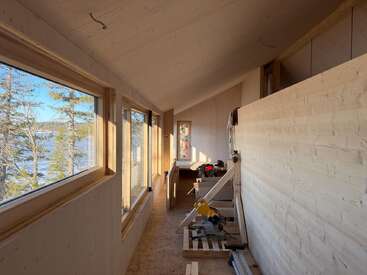This image shows a narrow, unfinished wooden room with large windows overlooking trees and a lake. Construction tools and materials are present, sunlight streaming in.