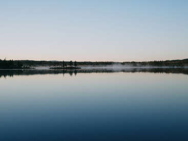 A calm, glassy lake reflects the clear sky above. Mist drifts gently over the water's surface, with a distant tree line silhouetted at sunrise.