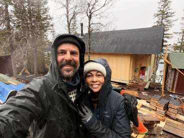 A smiling couple dressed in warm, outdoor clothing takes a selfie in front of a cabin under construction, surrounded by trees and building materials on a misty day.