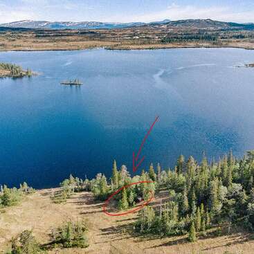 A serene lake surrounded by forest and mountains. A red arrow points to a small cabin hidden among trees near the shoreline, emphasizing its secluded location.